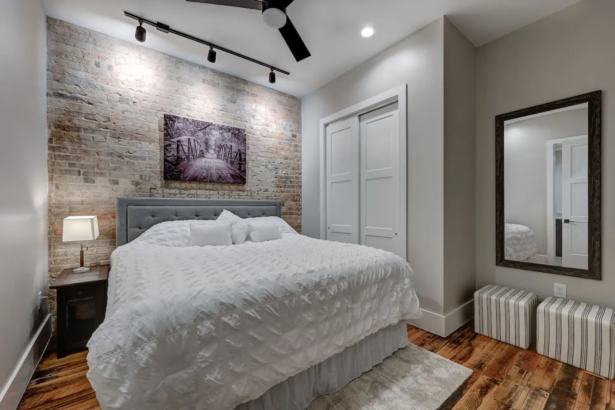 This image features a bed with crisp white bedding set against a striking exposed brick wall, accented by tasteful artwork and a stylish mirror. A nightstand with a glowing lamp adds a sense of warmth and intimacy, while the high ceiling amplifies the feeling of space.