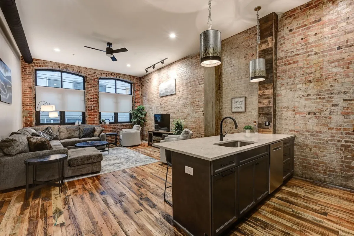 A spacious, open-concept living, dining, and kitchen area featuring a comfortable sofa, a stylish kitchen island, large windows that flood the room with natural light, exposed brick walls, and a television. The space seamlessly connects areas for lounging, cooking, and socializing, with contemporary finishes complementing the historic elements.