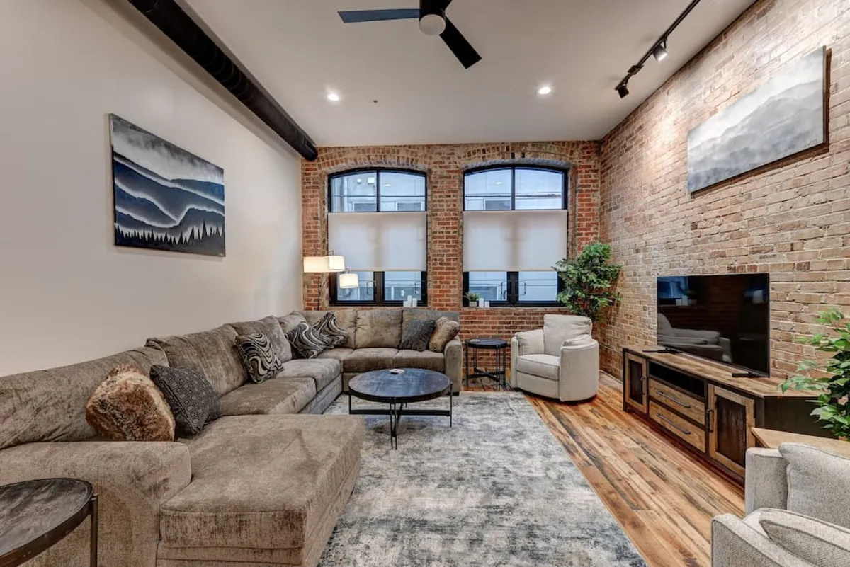 A stylish living room featuring a comfortable sofa, a modern coffee table, vibrant artwork on the wall, lush green plants, and a flat-screen TV. The space exudes a cozy, welcoming vibe, ideal for relaxing after a day of urban exploration or gathering with friends for intimate conversations.