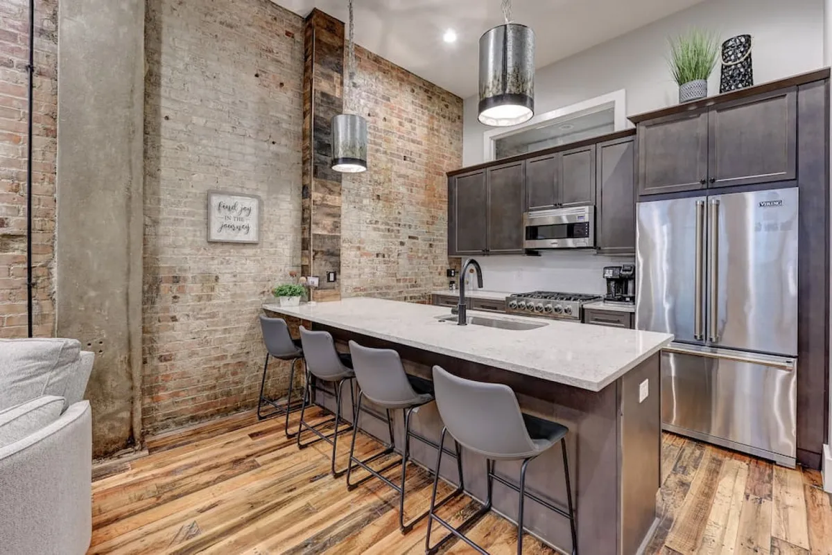 A modern kitchen highlighted by a prominent kitchen island with pendant lighting, a striking exposed brick wall, and stainless steel appliances. The layout is ideal for cooking and entertaining, with a stylish yet functional ambiance that invites both culinary creativity and casual gatherings.