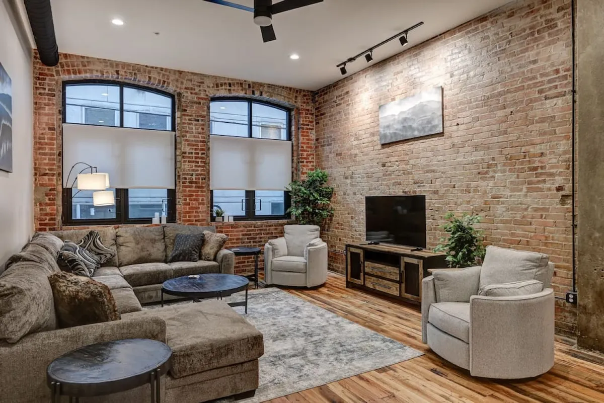 The hero image showcases a spacious, stylish living room with an exposed brick wall, a plush large sectional sofa, oversized windows flooding the room with natural light, a modern TV unit, and vibrant decorative plants. The space balances modern comfort with historic charm, inviting guests to unwind, socialize, or relax after exploring Asheville.