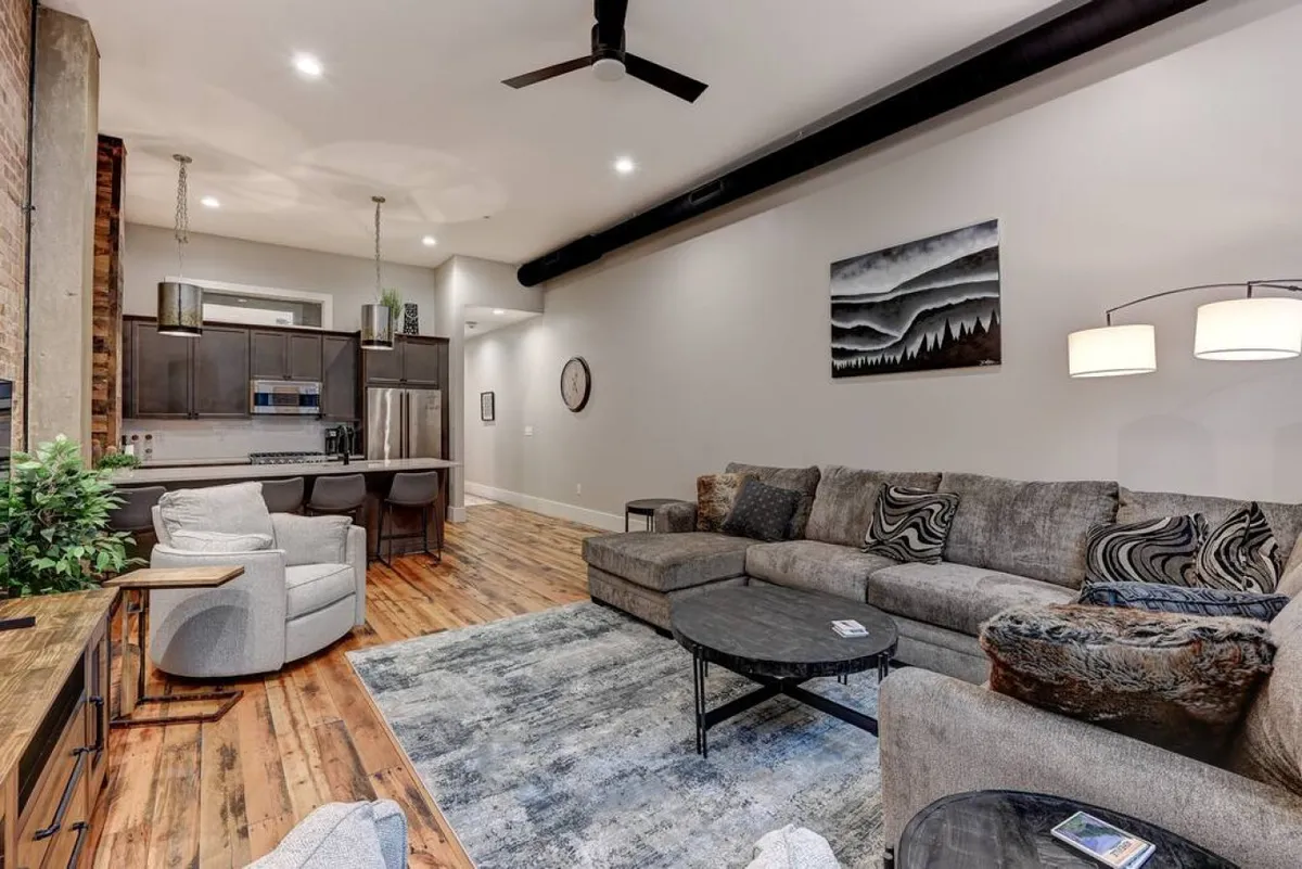 A comfortable and modern living area featuring a large sectional sofa, a central coffee table for gatherings, visible kitchen area in the background, and curated artwork on the wall. The space is designed for both relaxation and entertaining, with a light and airy feel that invites guests to linger.
